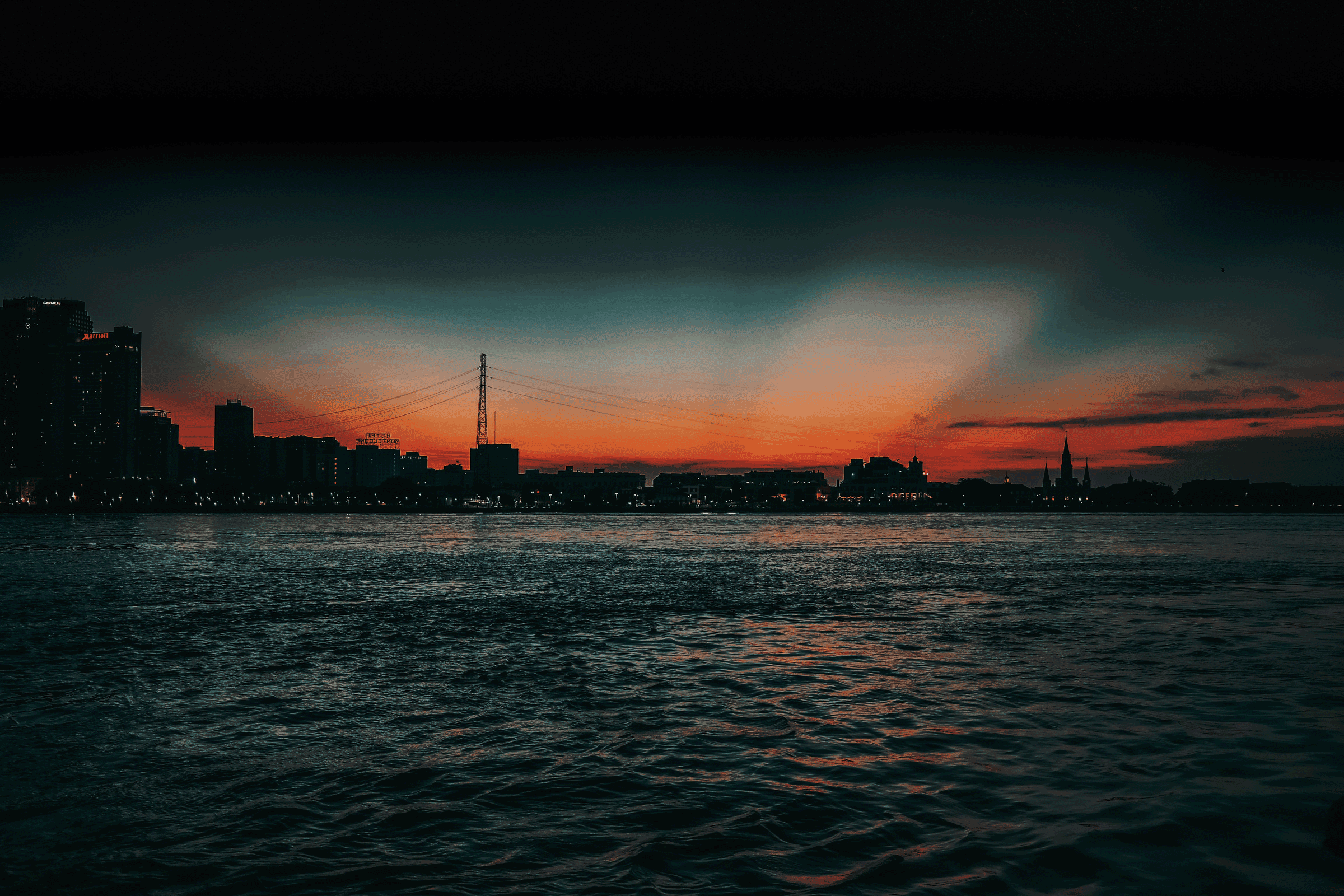 A sunset over a city skyline with water in the foreground, featuring tall buildings and a bridge.